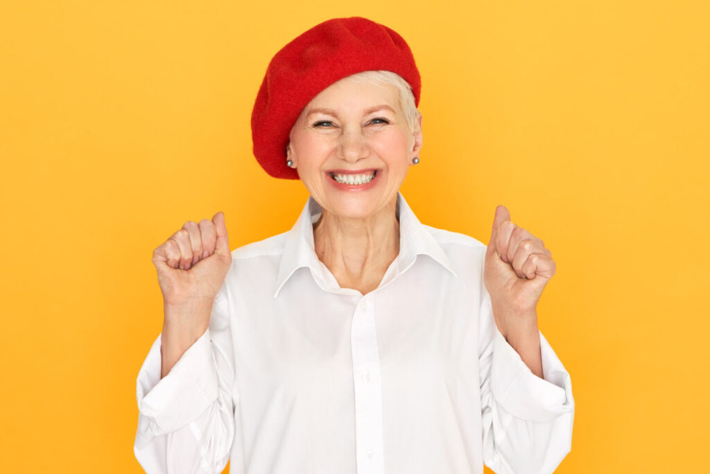 Idosa sorridente de boina vermelha celebra o rejuvenescimento facial e volume dos lábios com prótese protocolo na Clínica Perrella em São Caetano do Sul.