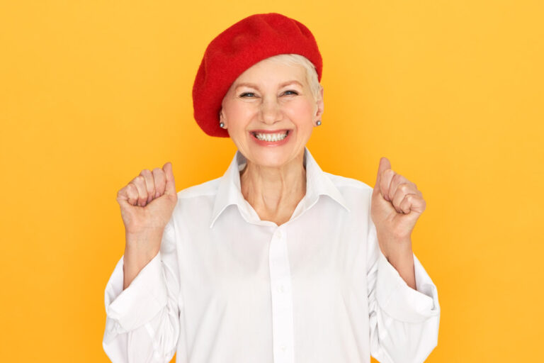 Idosa sorridente de boina vermelha celebra o rejuvenescimento facial e volume dos lábios com prótese protocolo na Clínica Perrella em São Caetano do Sul.