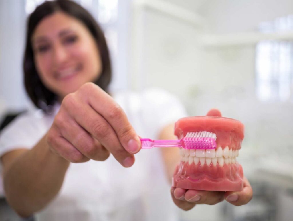 Dentista sorridente ensina a higienizar prótese protocolo usando escova e modelo anatômico na Clínica Perrella em São Caetano do Sul.